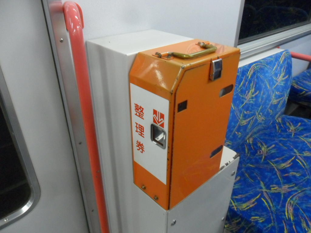 Gros plan d'un distributeur orange de tickets numérotés 整理券 (seiri-ken) monté à la porte d'un train Meitetsu au Japon